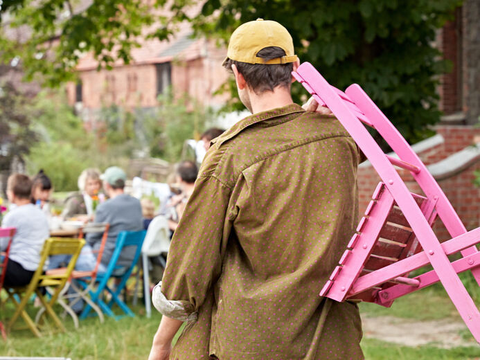 Ein Mensch trägt einen pinkfarbenen Klappstuhl zu einem gedeckten Tisch mit Menschen.